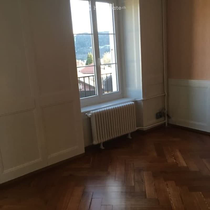 Empty room with wooden flooring, large window offering a view, radiator below the window, and neutral-colored walls.