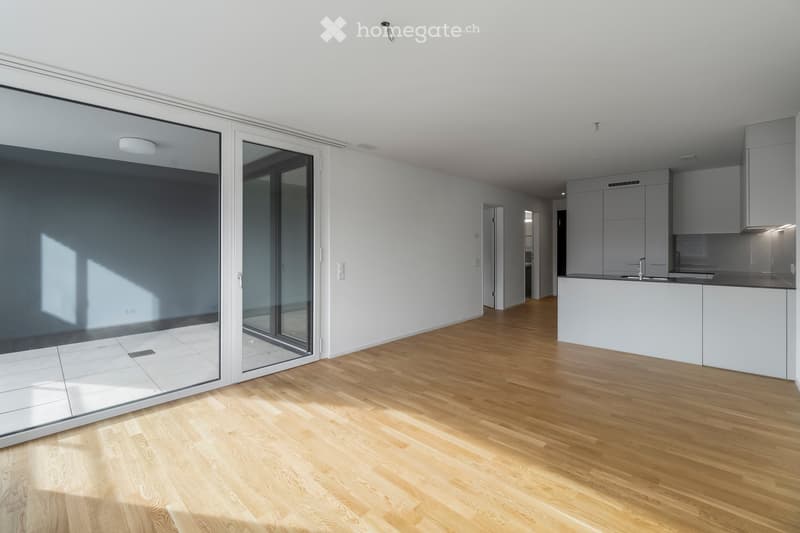 Bright, empty room with wooden flooring, large glass sliding doors, and a modern kitchen area visible in the background.
