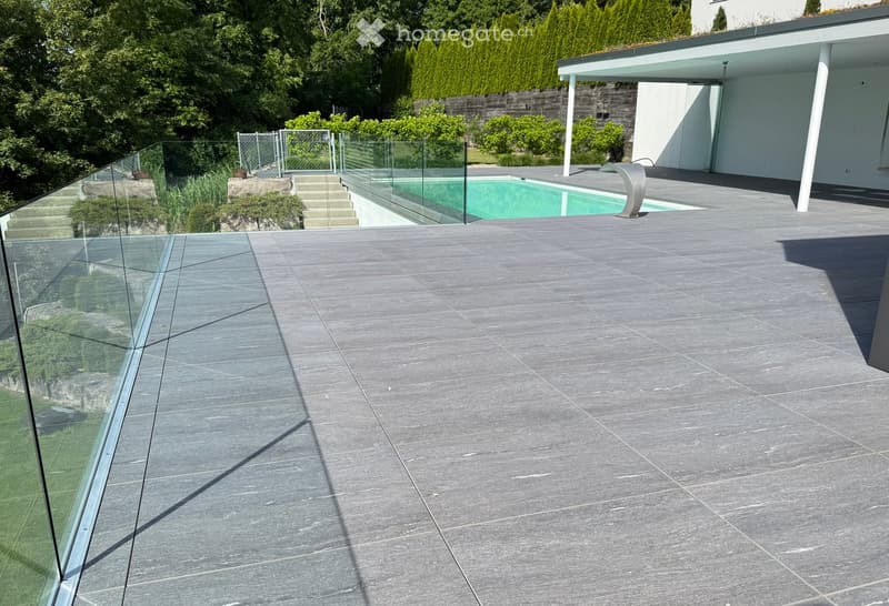 Modern outdoor patio with grey stone tiles, glass railing, swimming pool, and surrounding greenery.