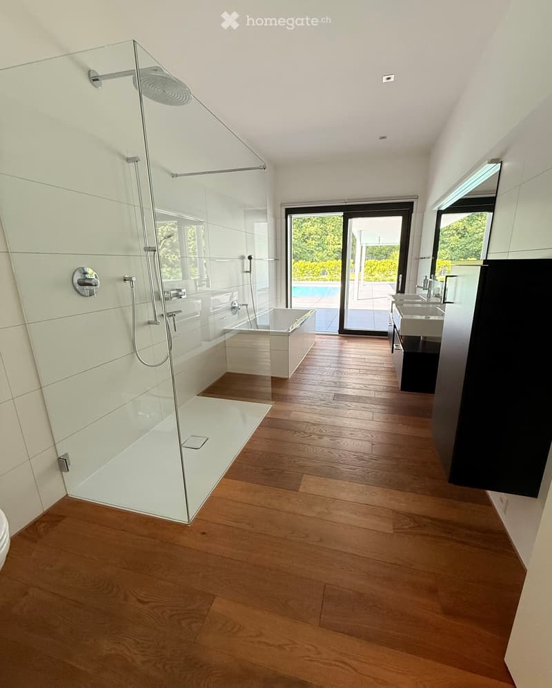 Modern bathroom with wooden flooring, a bathtub, a glass-enclosed shower, and a sink. A sliding glass door leads to an outdoor area with a swimming pool.