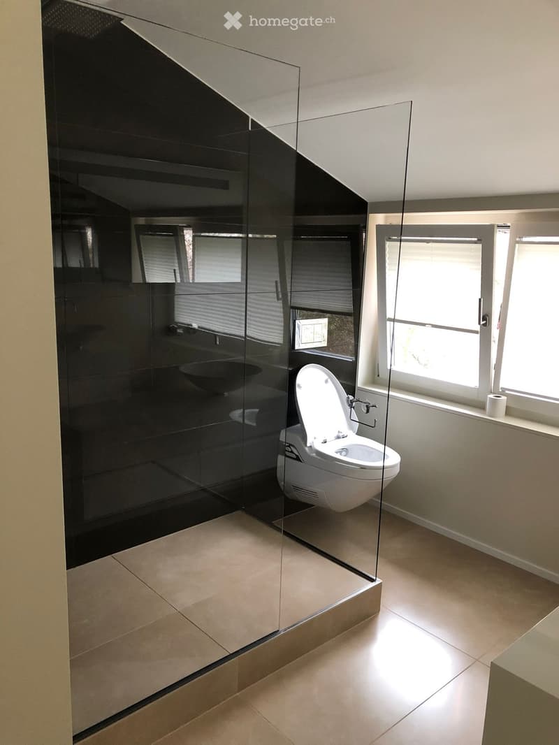 Modern bathroom with a toilet, sink, and large glass shower enclosure. Light-colored tiles and neutral walls.