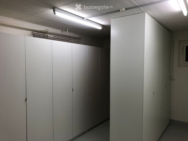 A narrow hallway with white walls, fluorescent lighting, and several white lockers lining one side. A metal basket is placed on top of the lockers.