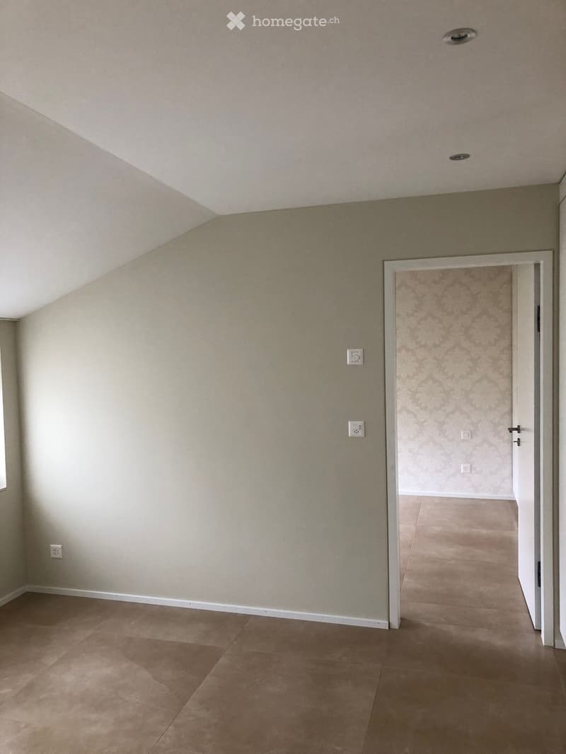 Empty room with beige walls, tiled floor, and a doorway leading to another room with patterned wallpaper.