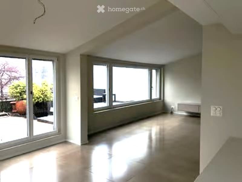 Bright, empty room with large windows and sliding glass doors leading to a balcony. The space features a polished concrete floor and white walls.