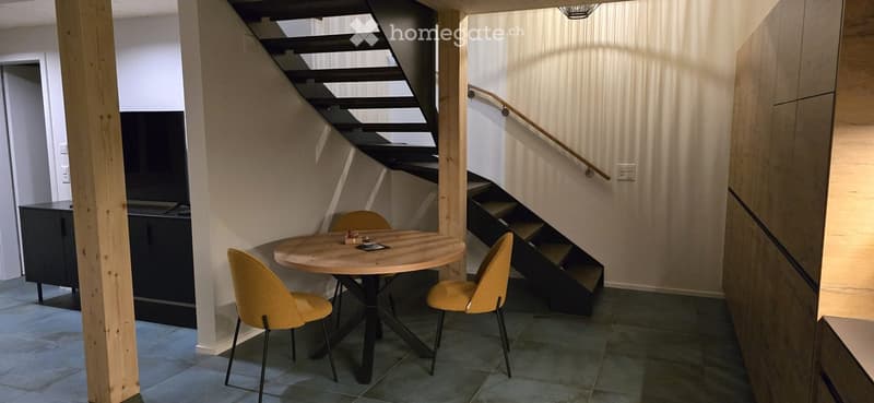 A modern, compact dining area with a round wooden table, four yellow chairs, and a sleek black cabinet. A staircase with a wooden railing is visible in the background.