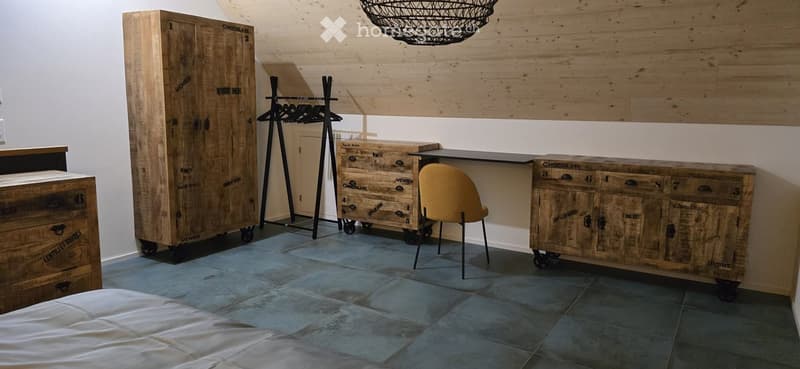 A stylish bedroom with a large wooden wardrobe, a wooden dresser, a desk, a yellow chair, and a wicker hanging lamp. The room features exposed wooden walls and a tiled floor.