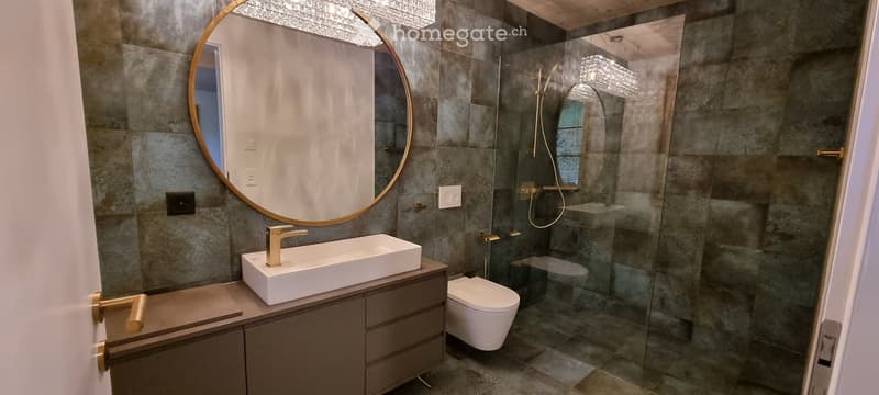 Modern bathroom with a white sink, gold faucet, large round mirror, glass-enclosed shower, and toilet.