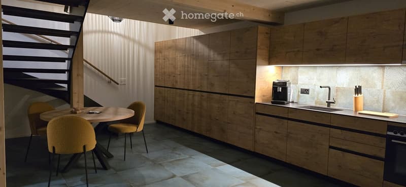 Modern kitchen with wooden cabinets, a round dining table with four chairs, a coffee maker, a knife block, and a cutting board on the counter.