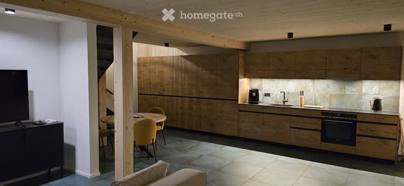 Open-plan living and kitchen area with modern wooden cabinets, a round dining table with chairs, a sofa, and various kitchen appliances including a coffee maker and teapot.