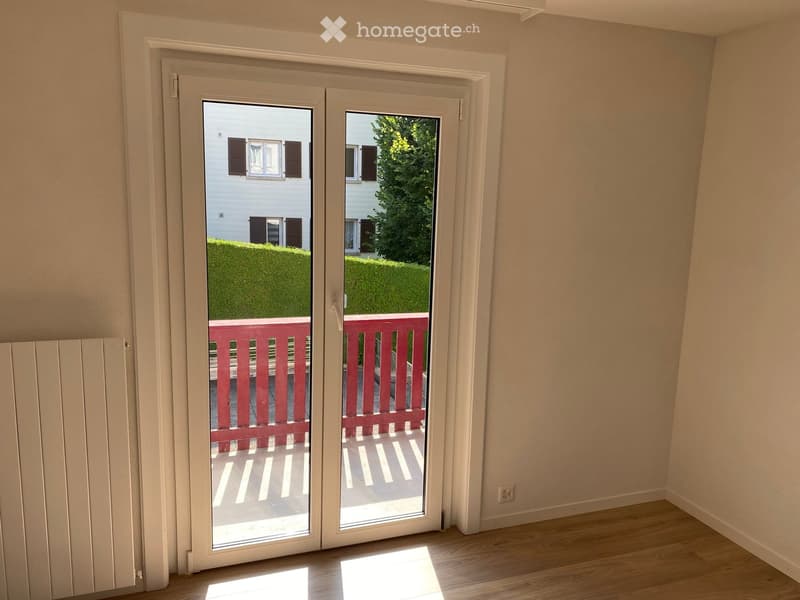 Empty room with light-colored walls and wooden flooring, featuring a large sliding glass door that opens to a balcony with a red railing and a view of a garden and neighboring house.