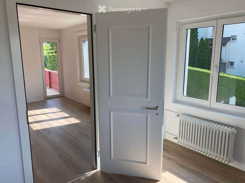Empty room with wooden flooring, white walls, and a large window with a view of greenery. Radiators are visible on both sides of the room.