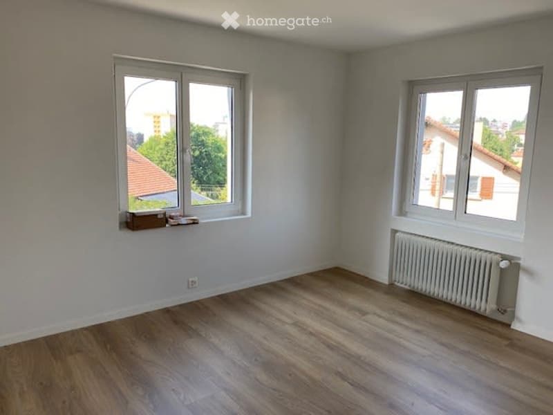 Empty room with white walls, wooden ceiling, and two windows offering views of the outside. Radiator visible on one wall.