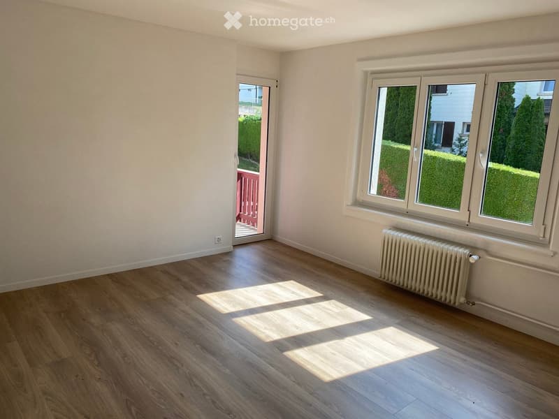Empty room with wooden flooring, white walls, large windows, and a radiator. A sliding door leads to an outdoor area.