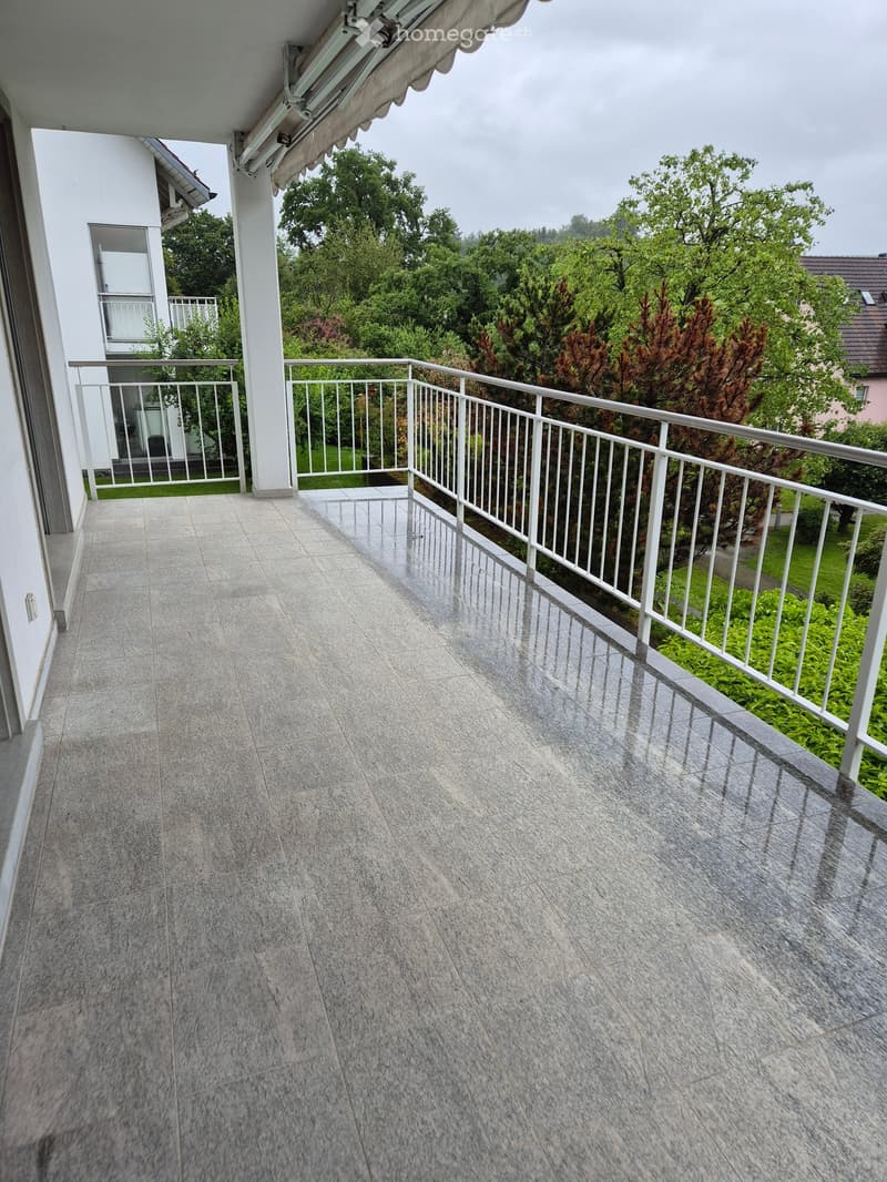 Balcony with tiled floor, white metal railing, and view of greenery and neighboring building.