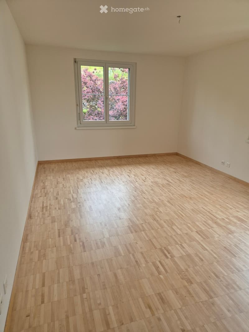 Empty room with wooden flooring and white walls, featuring a large window with a view of pink blossoms outside.