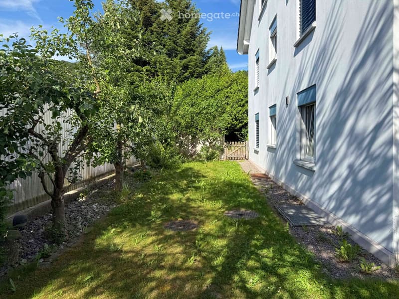 Garden area with grass, small trees, and a pathway leading to a white house with multiple windows.