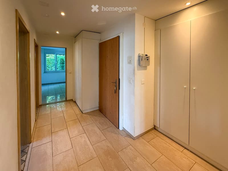 Clean hallway with tiled flooring, white walls, built-in storage cabinets, and recessed lighting. A wooden door leads to another room with a window showing greenery outside.