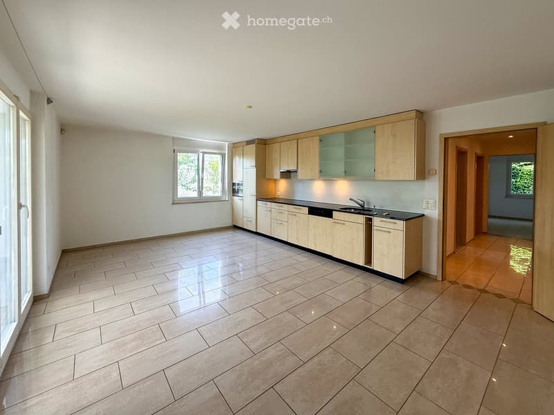 Empty kitchen with light wooden cabinets, tiled floor, and large windows allowing natural light. No appliances or furniture are present.