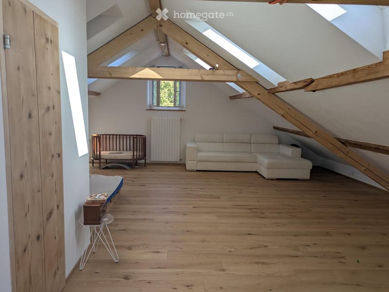Bright attic room with wooden beams, skylights, and wooden flooring. Features a cream-colored sectional sofa, a wooden baby crib, a small glass table with a stool, and a radiator.