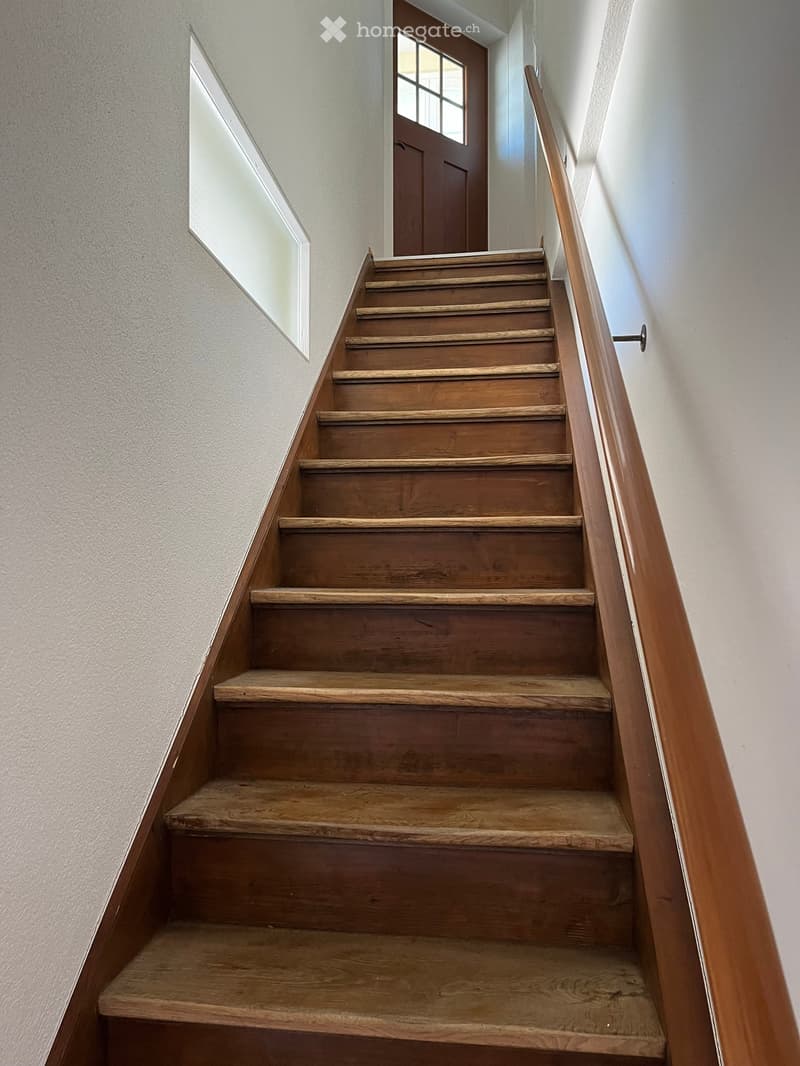 Wooden staircase with a wooden handrail, leading to a door with a window at the top of the stairs.