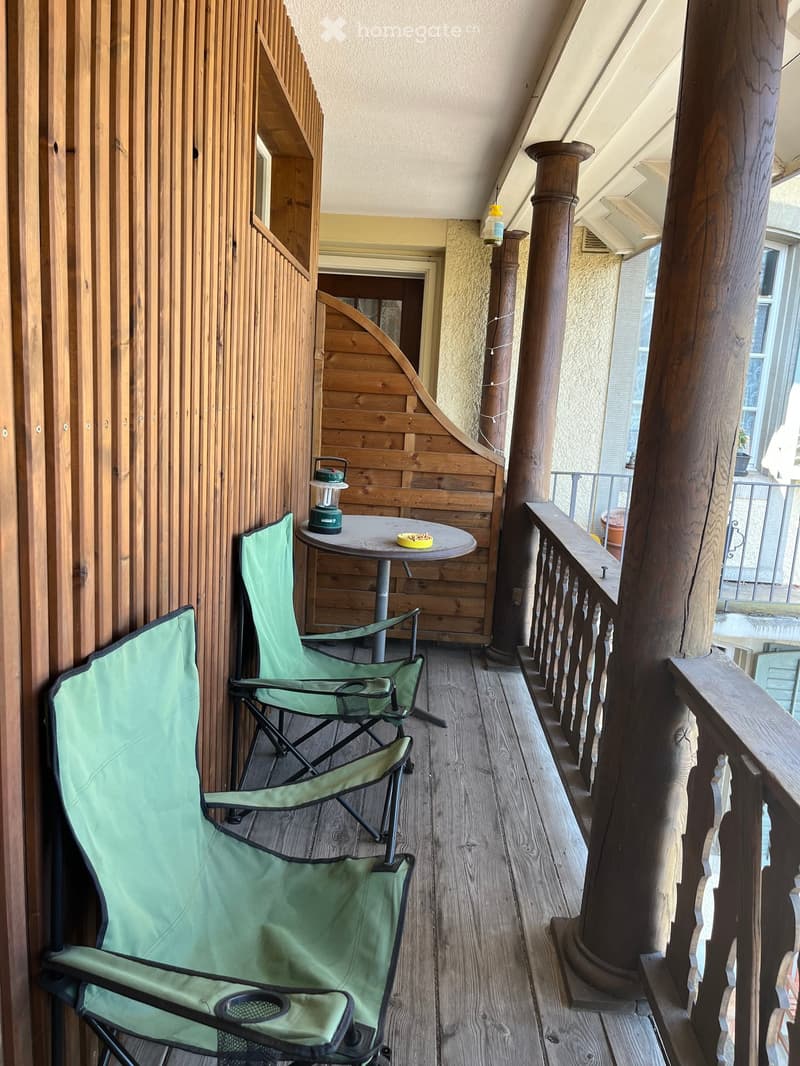 Balcony with wooden floor, two green folding chairs, small round table, lantern, and railing with wooden balusters.