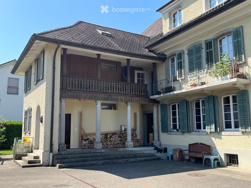 Two-story house with beige exterior, green shutters, and a balcony on the upper floor. Features include a wooden bench, small table, and stacked firewood near the entrance.