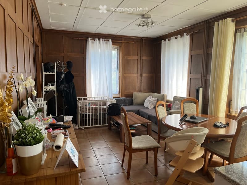 A well-lit living room with wooden paneling, a grey sofa, a wooden coffee table, a dining table with chairs, a baby chair, and a baby gate. There are decorative items including a flower vase and picture frames on a side table.