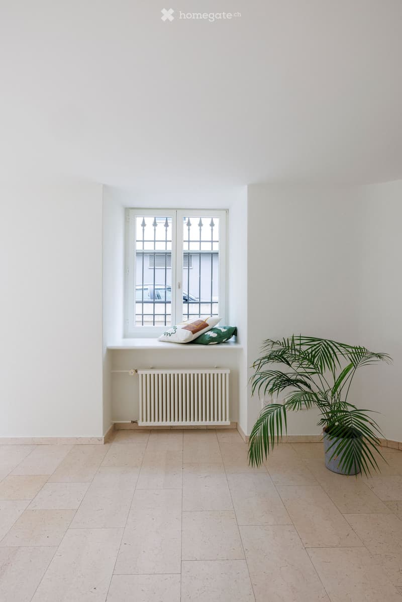 Empty room with light-colored tiled flooring, white walls, a window with decorative bars, a radiator, and a potted plant in the corner.