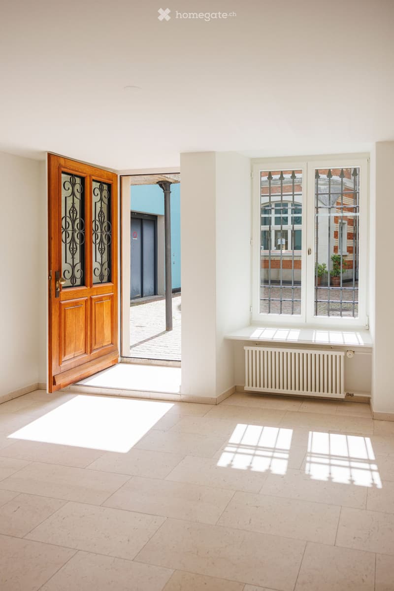 Empty hallway with tiled floor, white walls, large wooden door with glass panels, and a window with metal bars and radiator below.
