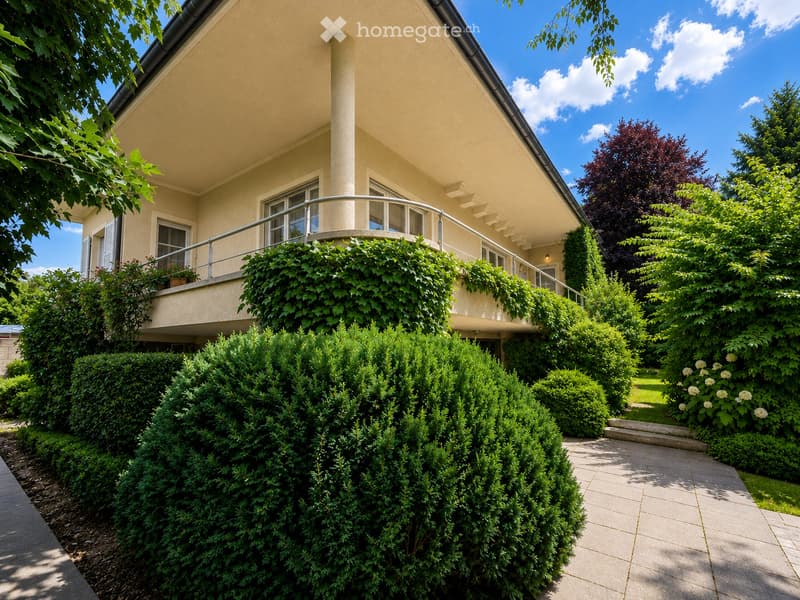 Two-story house with beige exterior, large windows, and a balcony covered in greenery. The garden features neatly trimmed hedges and a paved pathway.