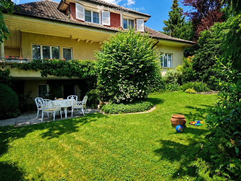 Lush garden with a well-maintained lawn, a patio area with a white table and chairs, and a large house with yellow and red exterior walls and multiple windows.