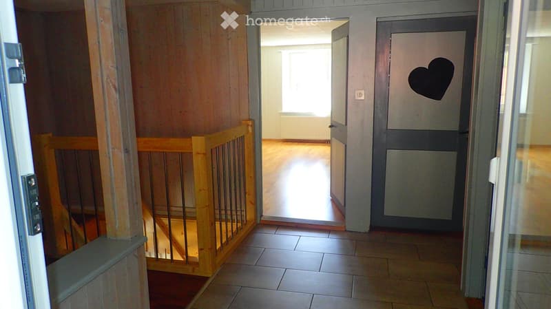 Hallway with tiled floor, wooden staircase railing, open doorway leading to another room with wooden flooring, and a heart-shaped sticker on the wall.