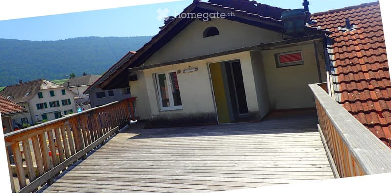 A two-story house with a wooden deck and railing, overlooking a scenic valley with mountains in the background.