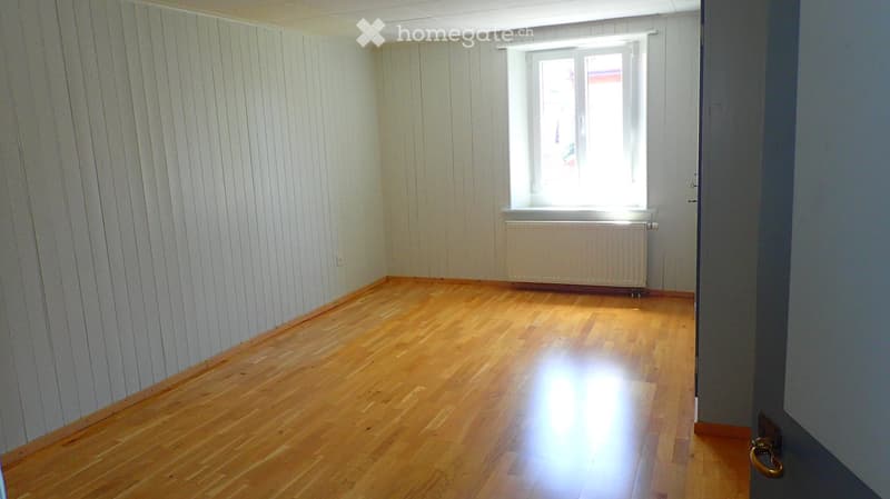 Empty room with wooden flooring, white paneled walls, a window with white frame, and a radiator below the window.