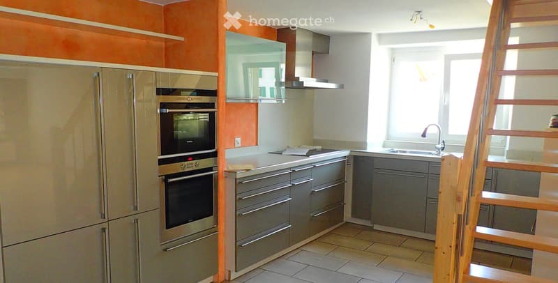 Modern kitchen with stainless steel appliances, including a refrigerator, oven, and stove, along with grey cabinets and a sink with a faucet. The space features a tiled floor and a wooden staircase leading upwards.