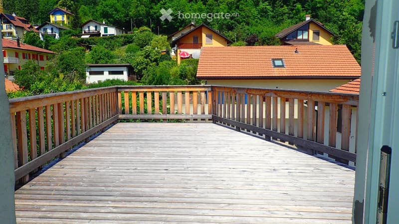 Wooden balcony with railing, overlooking a residential area with houses and greenery.