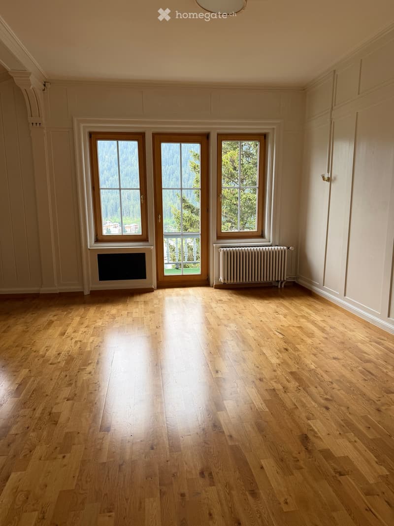 Empty room with light wooden flooring, three large windows with wooden frames, a fireplace, and a radiator. The walls are painted in a light cream color.