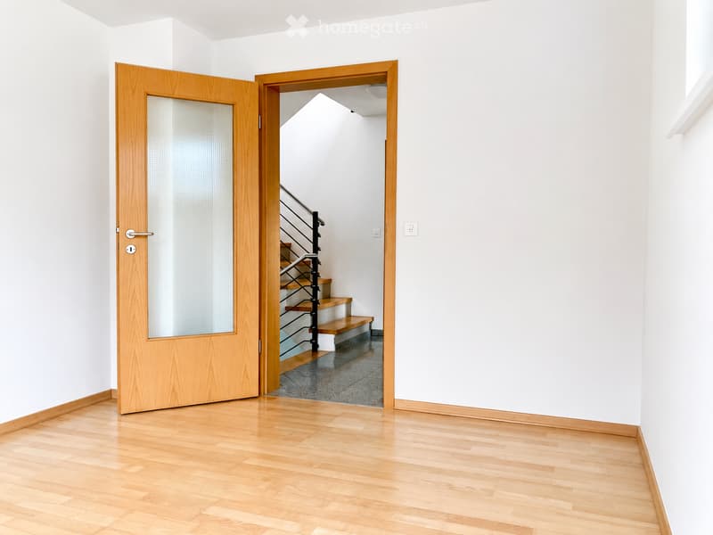 Empty hallway with light wooden flooring and white walls, featuring an open wooden door leading to a staircase with a metal railing.