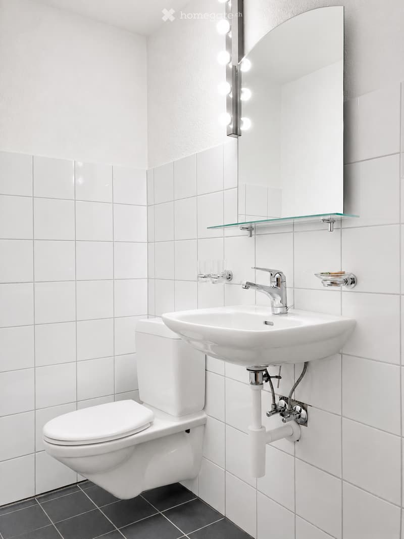 Modern bathroom with white tiles, a toilet, a sink with a modern faucet, a large mirror with built-in lighting, and a glass shelf above the sink.