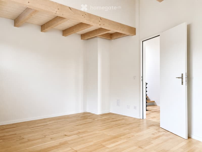 Empty room with light wooden flooring, white walls, and exposed wooden beams on the ceiling. A white door is open, revealing a glimpse of another room with a staircase.