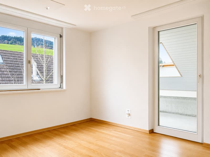 Empty room with light-colored walls, wooden flooring, and large windows offering a view of the outdoors.