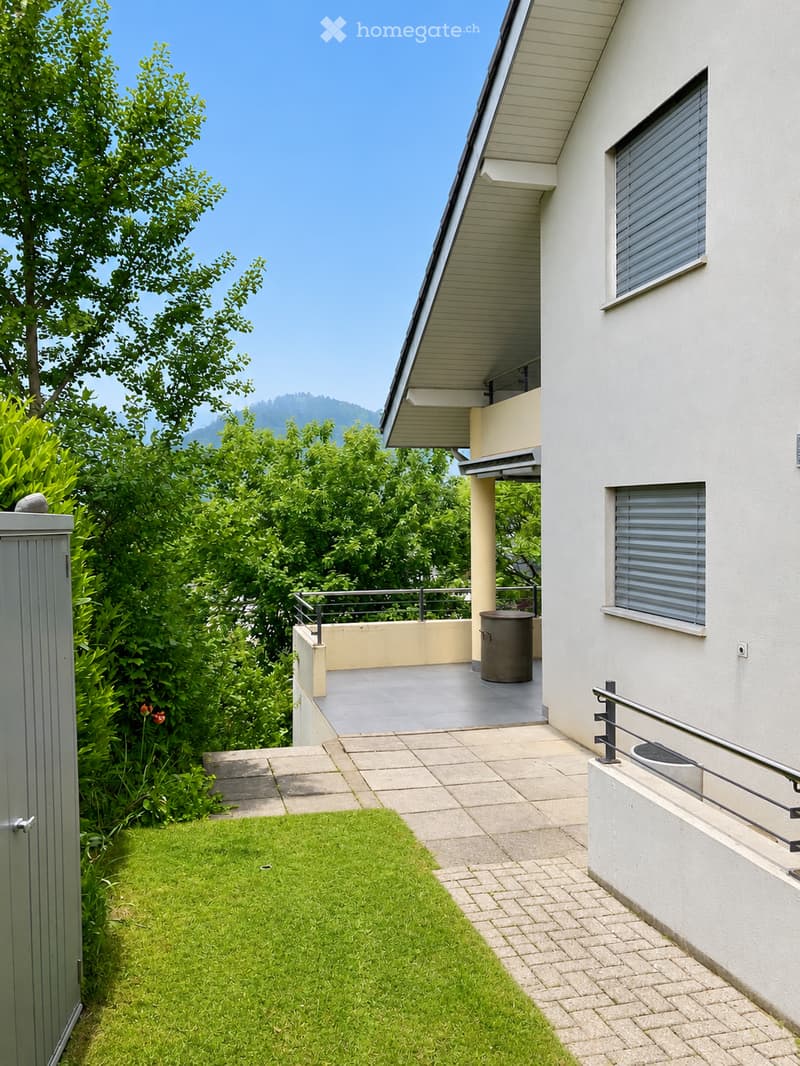 Modern house exterior with a well-maintained garden, a paved pathway, and a small balcony area with a railing and a trash bin.