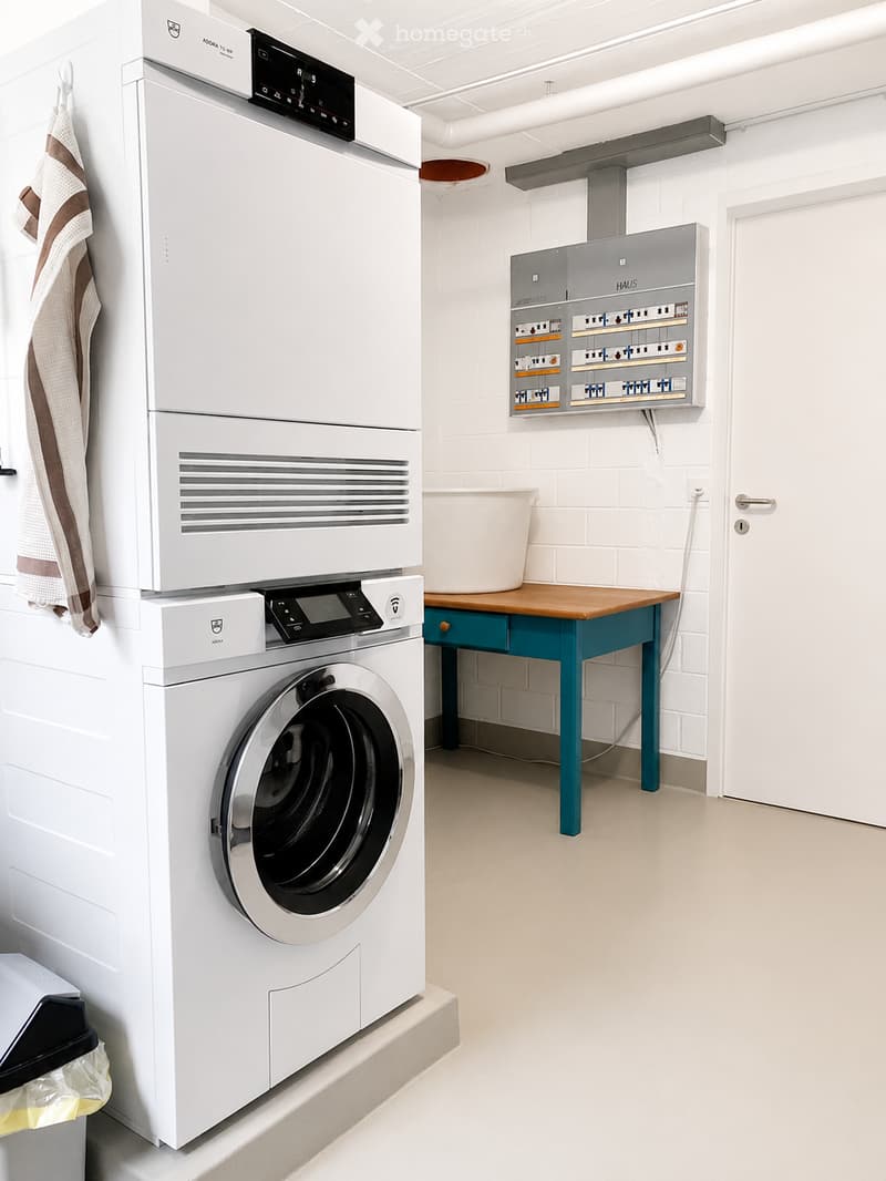 Modern laundry room with white washing machine, drying cabinet, small wooden table with stool, electrical panel, and a hanging towel.