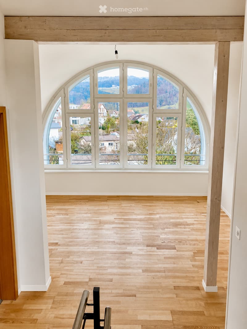 Bright, empty room with wooden flooring and large arched windows offering a scenic view of the surrounding landscape.