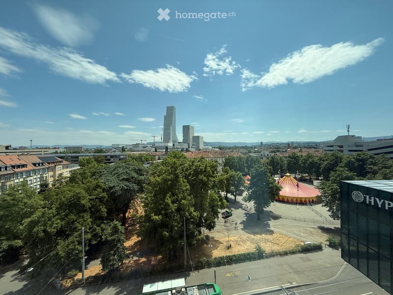 Aerial view of a cityscape with modern high-rise buildings, a circus tent, trees, and a paved area with a few people and vehicles.