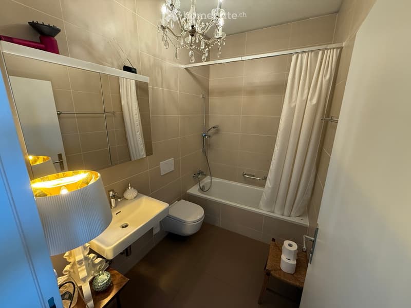 Modern bathroom with beige tiles, a bathtub, toilet, sink, mirror, chandelier, and a small table with a lamp and decorative items.
