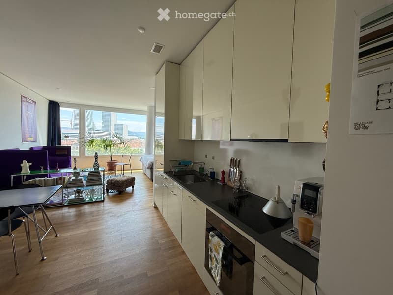 Open-plan living and kitchen area with modern white cabinets, black countertops, and a sleek coffee maker. The living area features a purple sofa, glass coffee table, and a small dining table with chairs. A potted plant adds a touch of greenery, and large