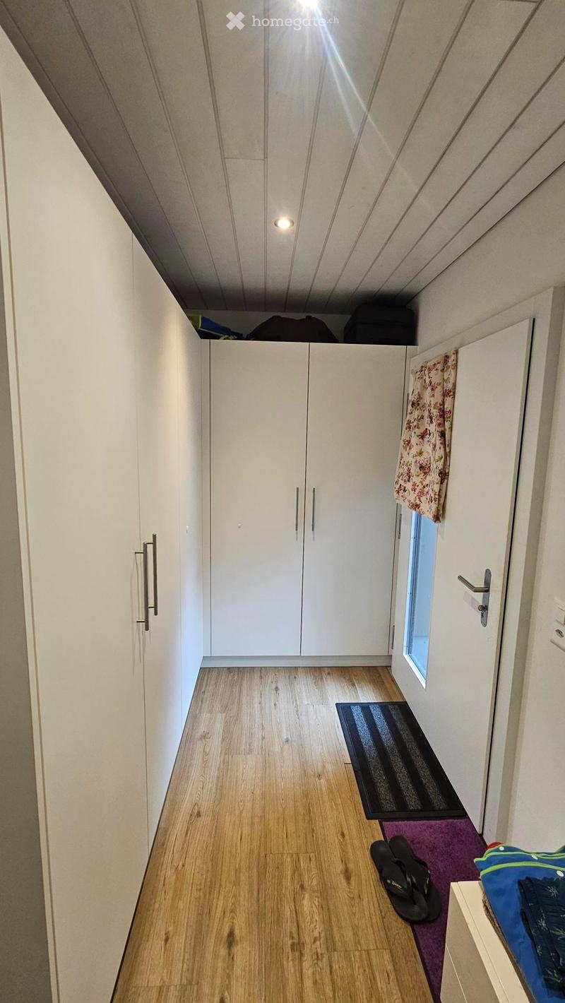 A narrow hallway with light wooden flooring, white walls, built-in white cabinets with silver handles, a small window, and a purple rug with shoes and a bag placed on it.