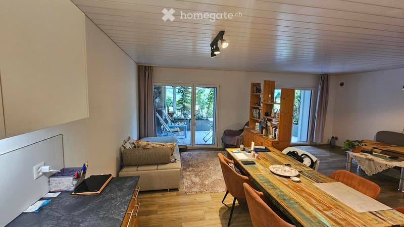 A modern, open-concept living and dining area with a sofa, dining table, chairs, bookshelf, and a small plant. The space features large windows and a sliding glass door leading to a balcony.