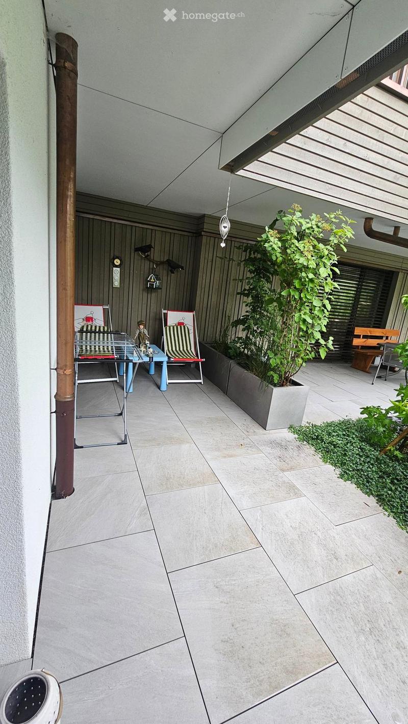 Modern outdoor patio with light-colored tiles, a small table with chairs, a potted plant, and a bench.